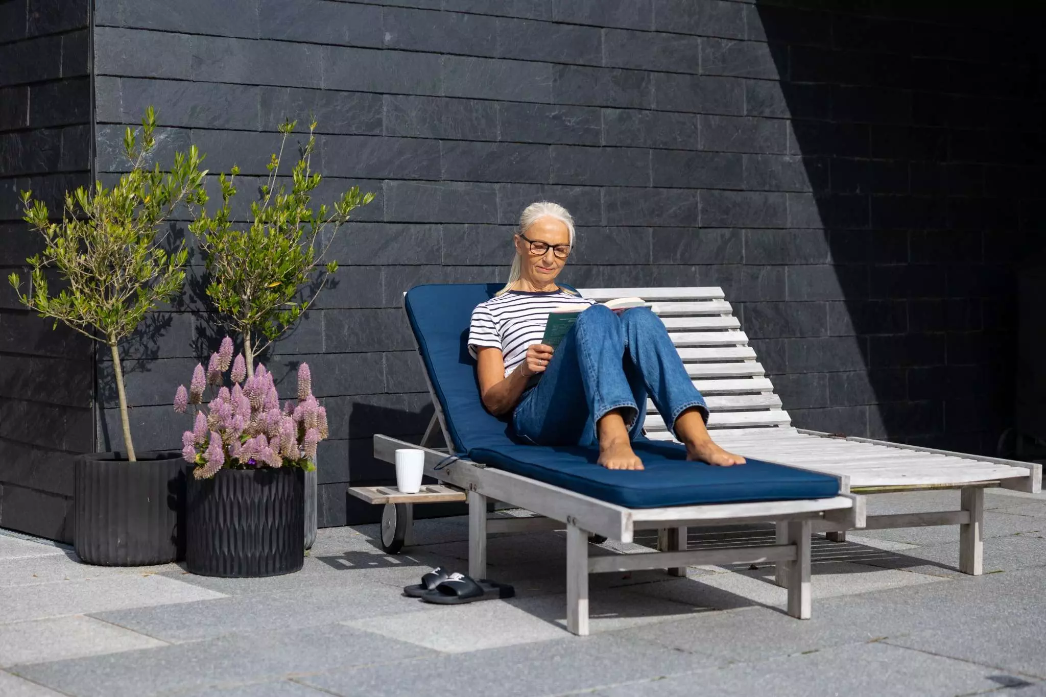 En kvinde med briller sidder på en liggestol med en blå hynde og læser en bog. Hun er iført en stribet t-shirt og jeans. Ved siden af liggestolen står en hvid kop og et par sandaler på en terrasse med fliser. To blomsterkrukker med planter står ved siden af en mørk murstensvæg.