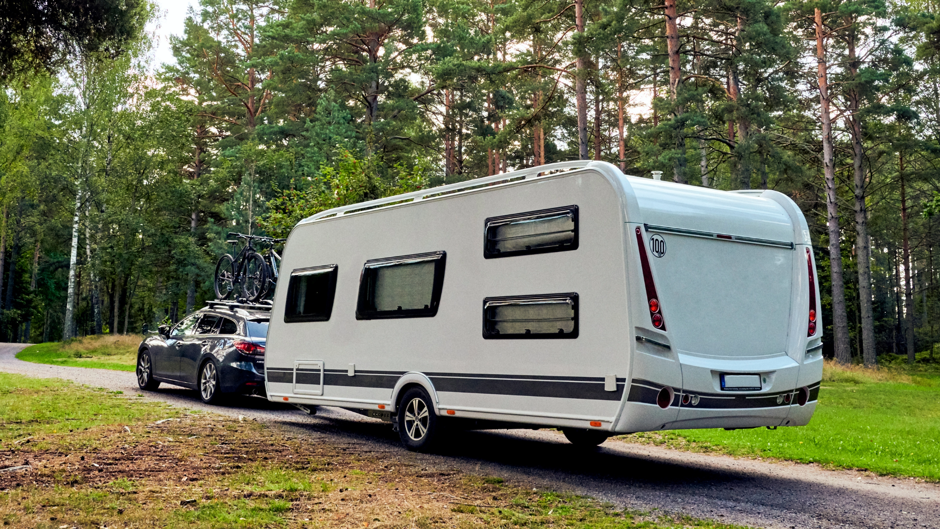 Bil med monterede cykler på taget trækker en moderne campingvogn ad en skovvej omgivet af høje træer.