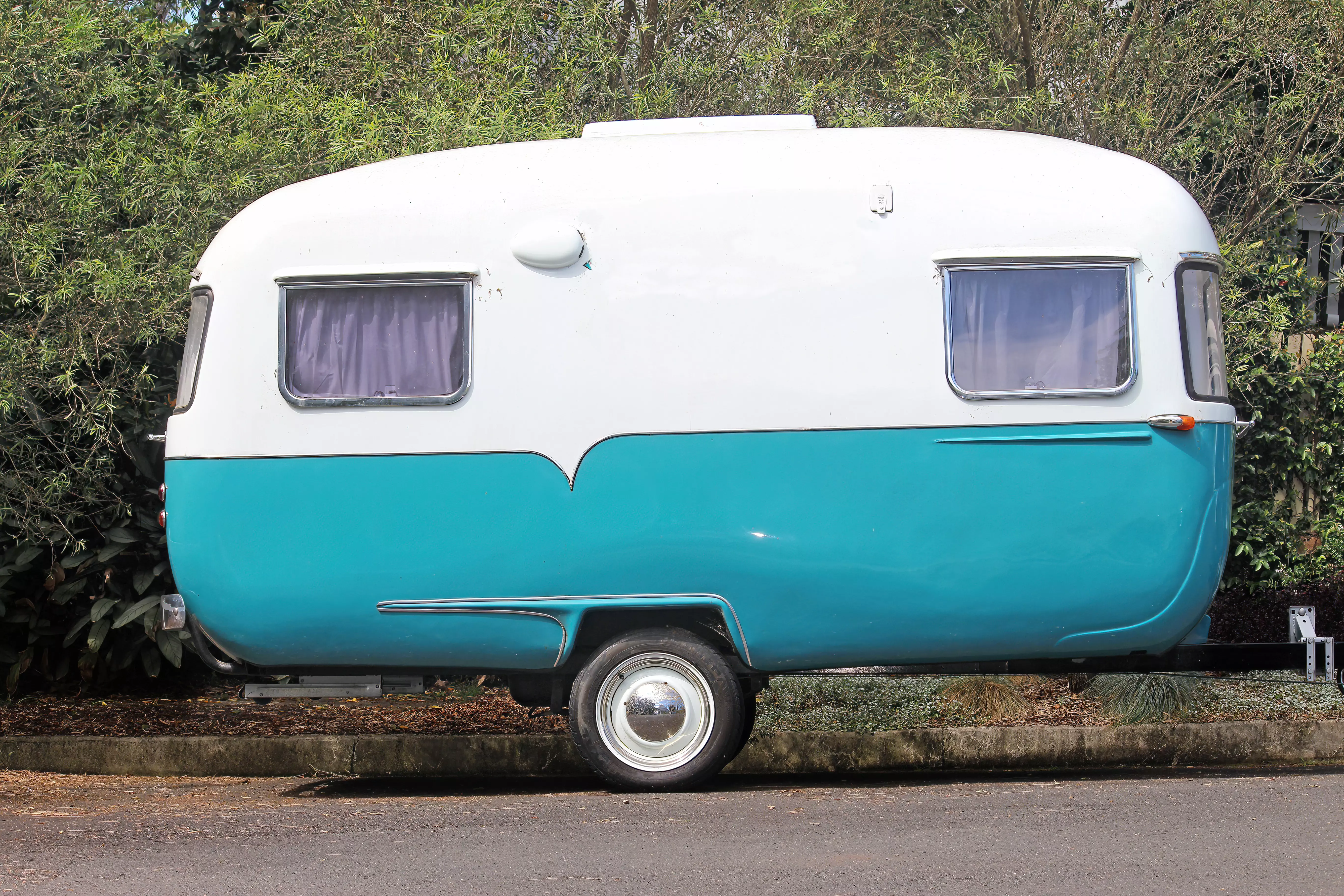 En lille retro campingvogn i to farver, hvid foroven og turkis forneden, parkeret på en asfaltvej med grøn beplantning i baggrunden. De små vinduer har gardiner trukket for, og det skinnende hjul reflekterer lyset.