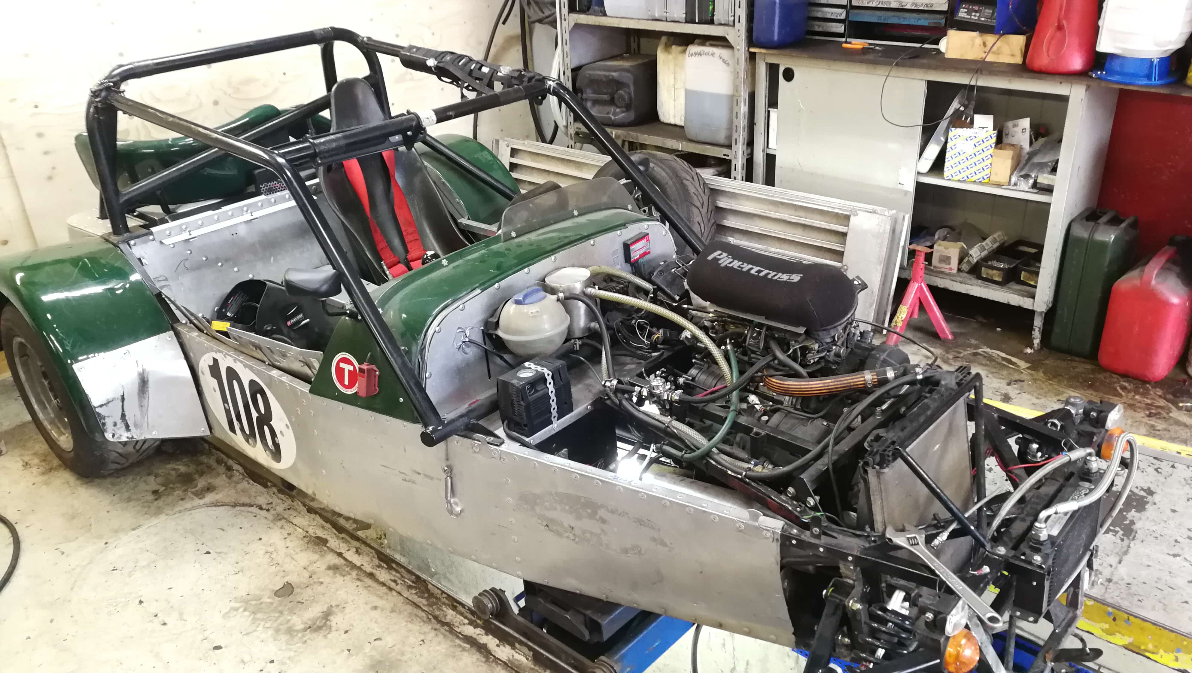 En ufuldendt racerbil i et værksted med et råt og åbent design. Bilen har et grønt og sølvfarvet karrosseri med nummer "108" på siden, en rørformet sikkerhedsbur og en synlig motor med rør og kabler. Der er to sæder med røde seler, og værkstedsbaggrunden indeholder værktøj, reservedele og benzin- eller oliedunke.