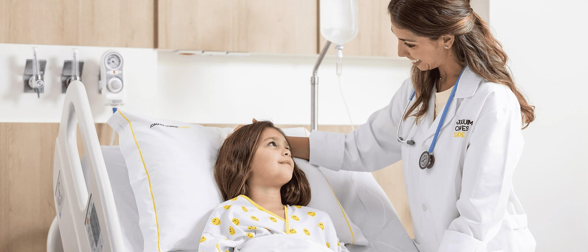 Médica pediátrica em pé e uma paciente numa cama de hospital