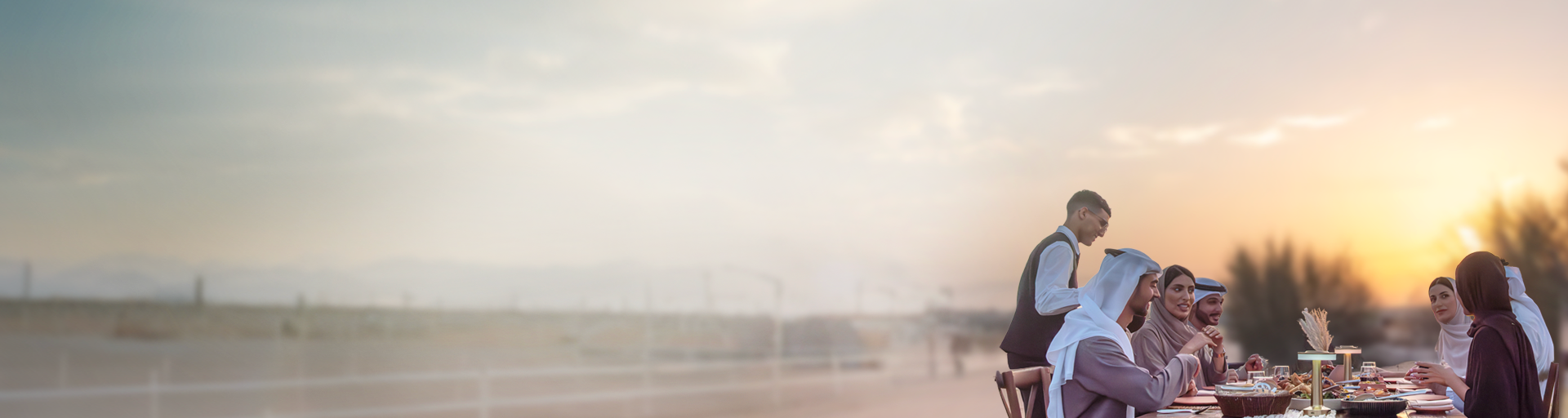 A family in UAE traditional clothing enjoying dinner outdoors, assisted by DWTC Hospitality staff.