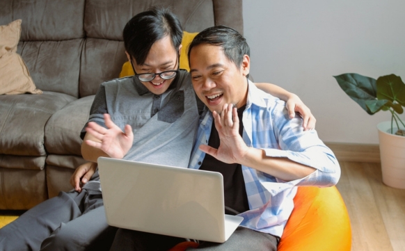 A young man and his father sit closely together waving and smiling at a laptop.