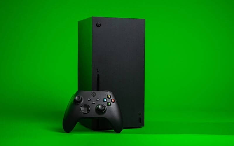 A black Xbox X console with a black Xbox controller leant against the console, with a bright green background.