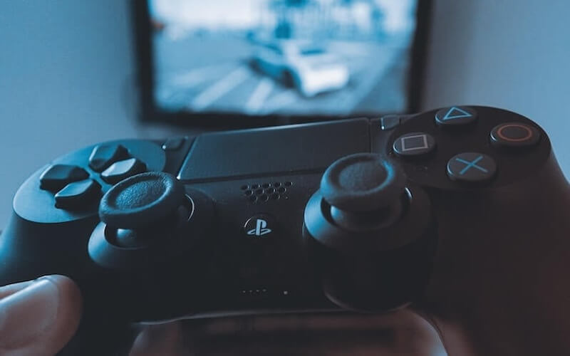 Hands holding a black PS4 controller with a TV screen in the background showing a car racing game on the screen.