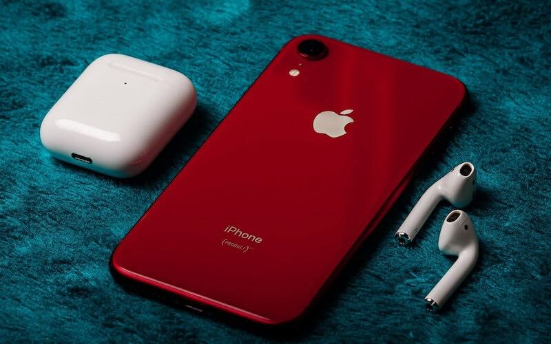 Red iPhone XR with white Apple Airpods case and earbuds on either side of the phone.