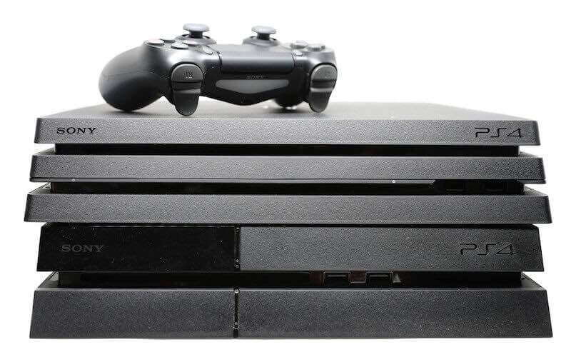 A black PS4 console and a black PS4 Pro console both lay on top of each other with a black PS4 controller sat on top of the two consoles.
