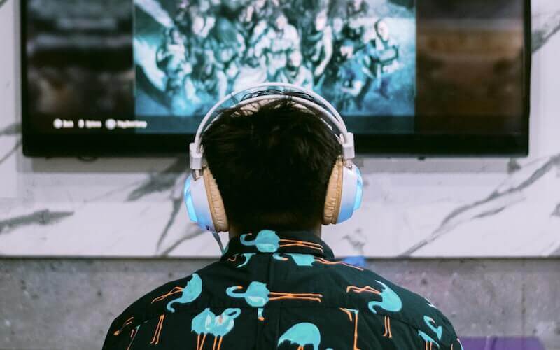 A man wearing white headphones and a patterened shirt is facing a larger TV screen while playing on a games console