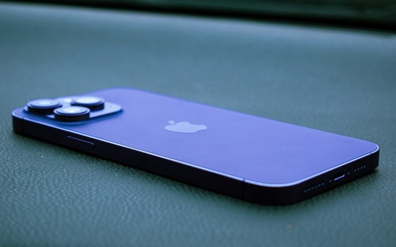 Purple iPhone Pro model lay on a leather surface with camera and back of device pointing upwards.