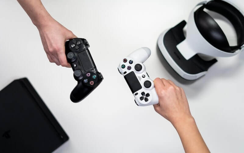 Two hands outstretched over a white surface: one hand holds a black PS4 controller and the other hand holds a white PS4 controller. There is a black PlayStation console and a VR headset also in shot.