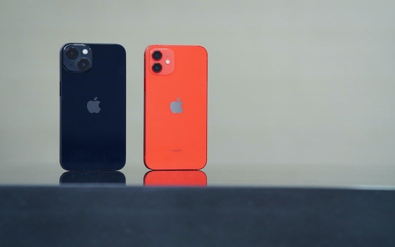 Two iPhone models stood up next to each other. One Dark coloured iPhone 13 and one bright red iPhone 12.