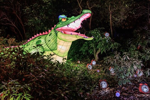Image of an outdoor area with a green and pink inflatable crocodile peeking out of the bushes, surrounded by a number of small red alarm clocks.