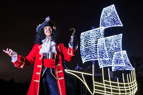 Image of a man dressed in a pirate costume with a red coat, black hat and a hook on one hand. The man is standing in front of a large pirate ship made from small lights.