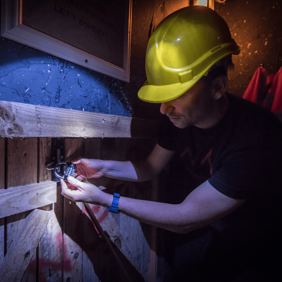 A man in a dark room wearing a hard hat focuses on unlocking a padlock with a 4 digit code