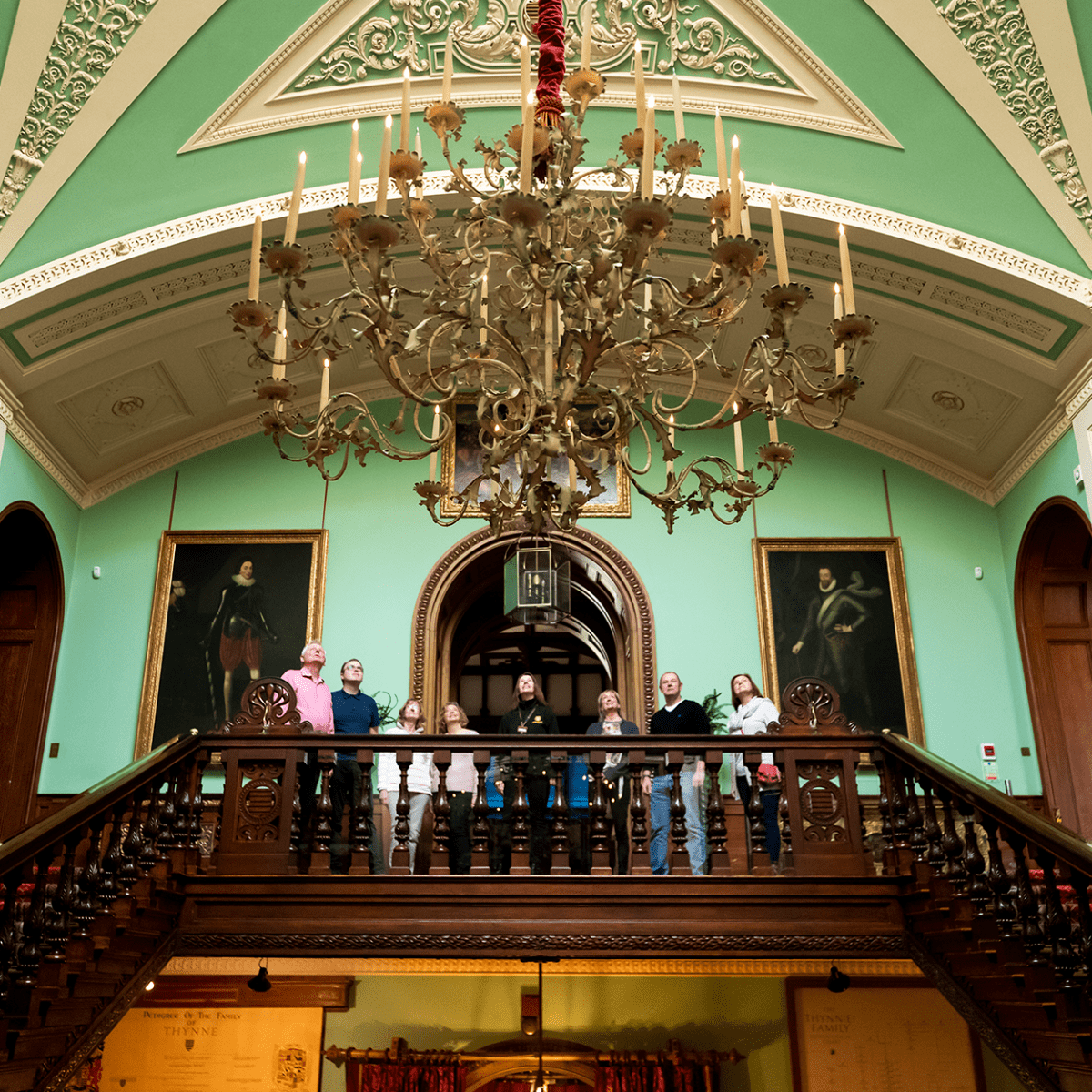 A group of visitors stand at the top of the staircase to look up at the grand chandelier on a guide tour of Longleat House