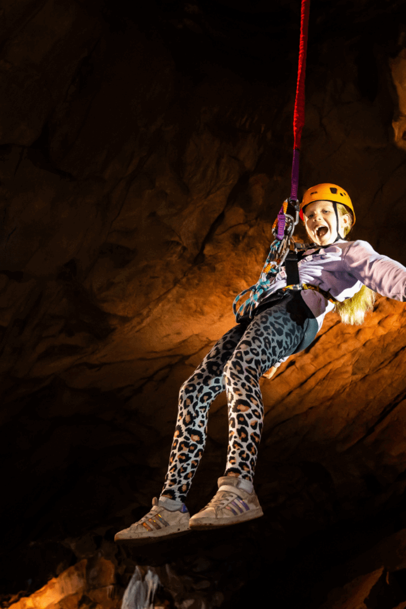 A young girl experiencing the Black Cat Freefall challenge with a bright smile on her face as she is suspended in the cave