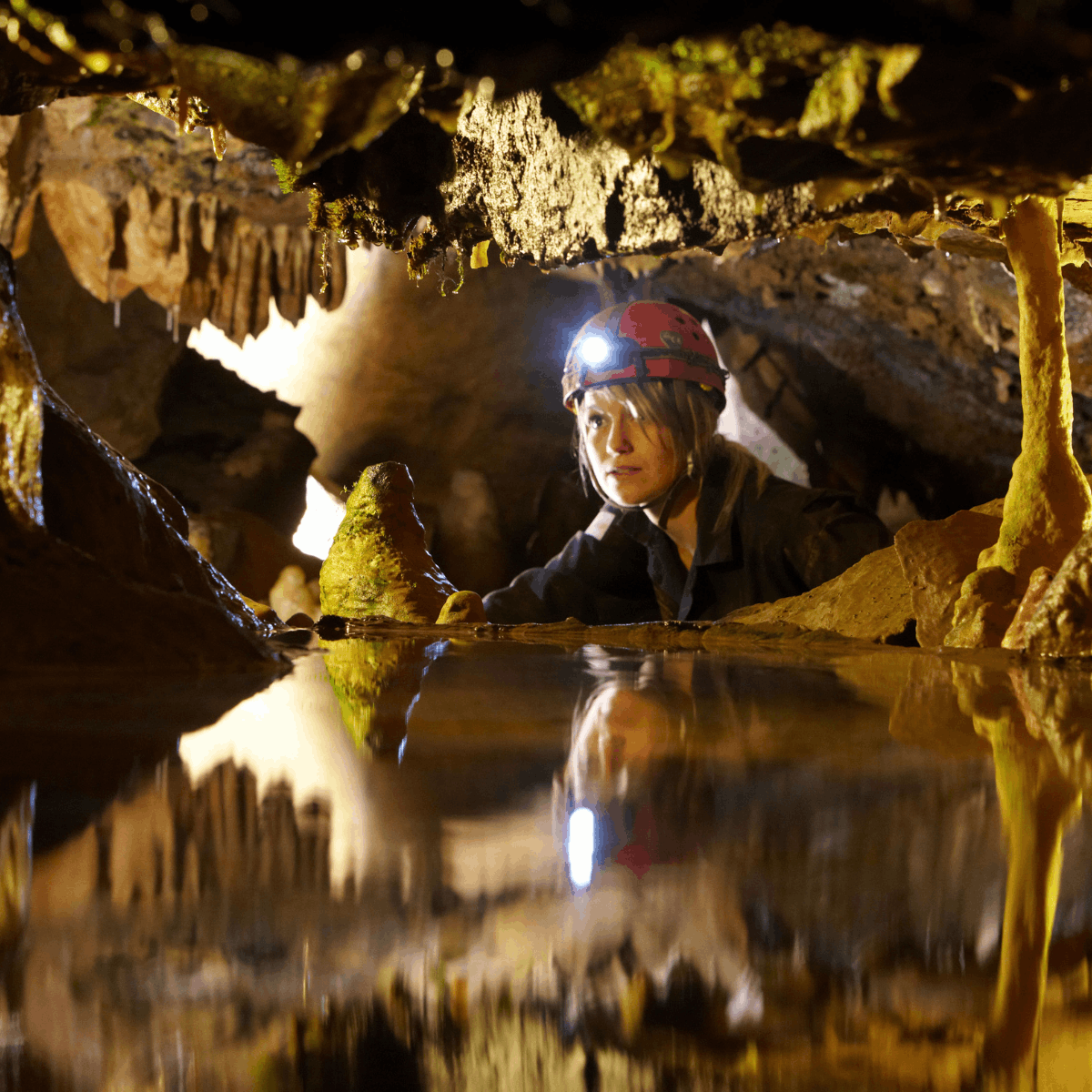 A woman wearing a helmet and head torch looks through a gap in rocks within a cave