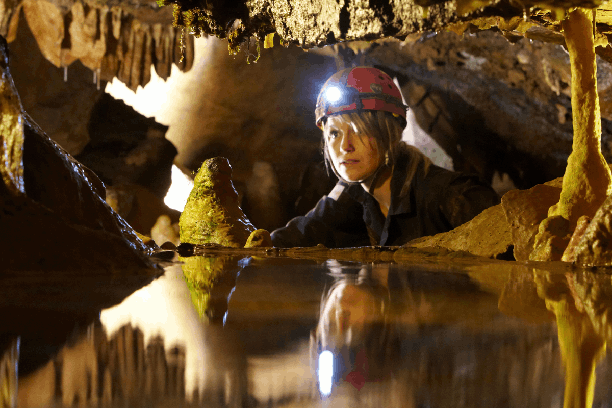 A woman wearing a helmet and head torch looks through a gap in rocks within a cave