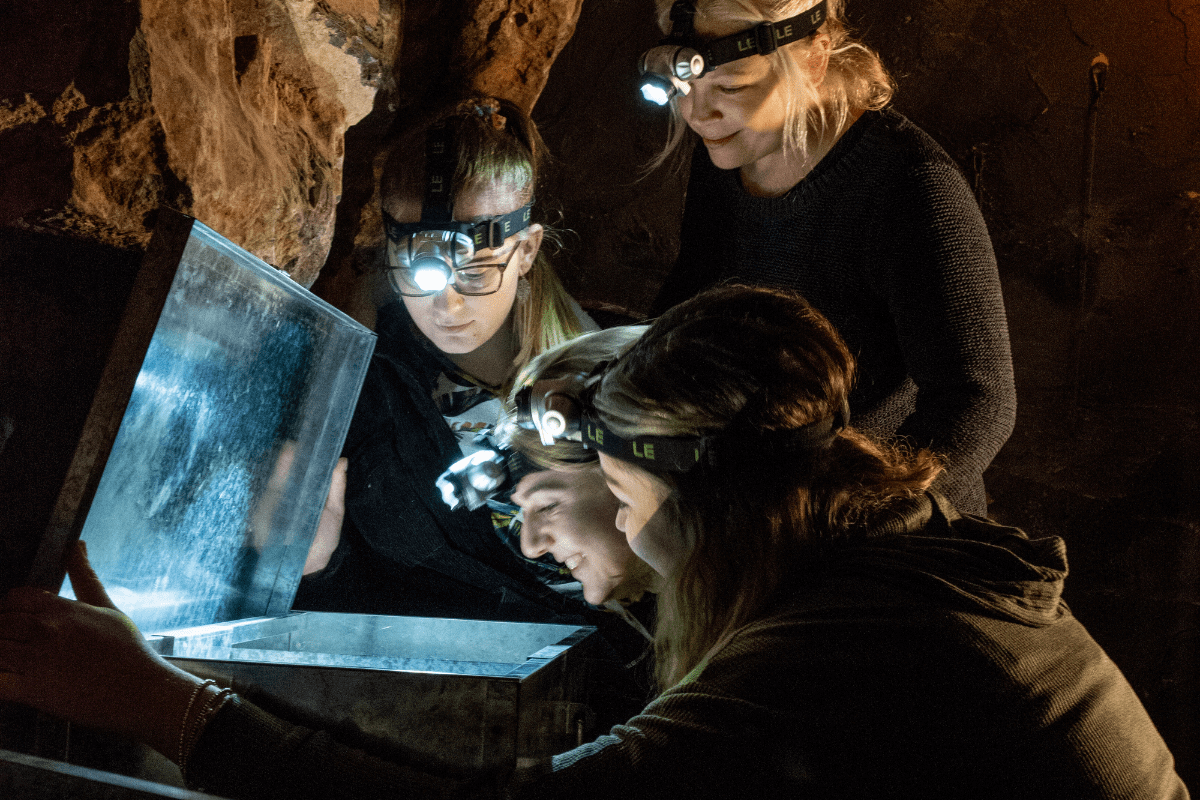 A group of people look at a potential clue in the escape rooms at Cheddar. They gather round a lit up chest looking for answers