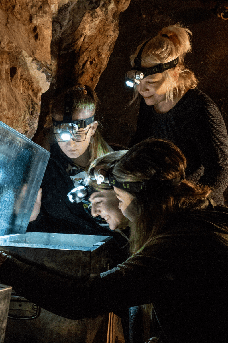 A group of people look at a potential clue in the escape rooms at Cheddar. They gather round a lit up chest looking for answers