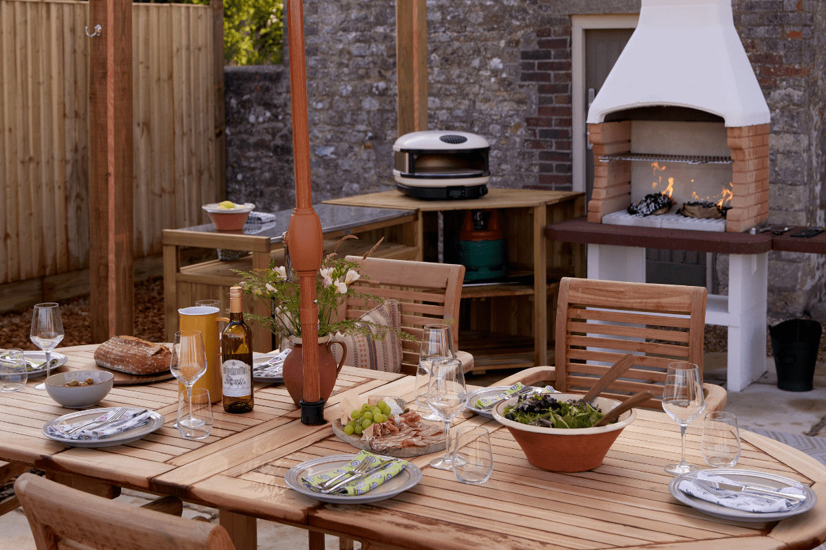 An outdoor dining area with wooden table and chairs, laid with plates and glasses in front of a lit pizza oven and grill