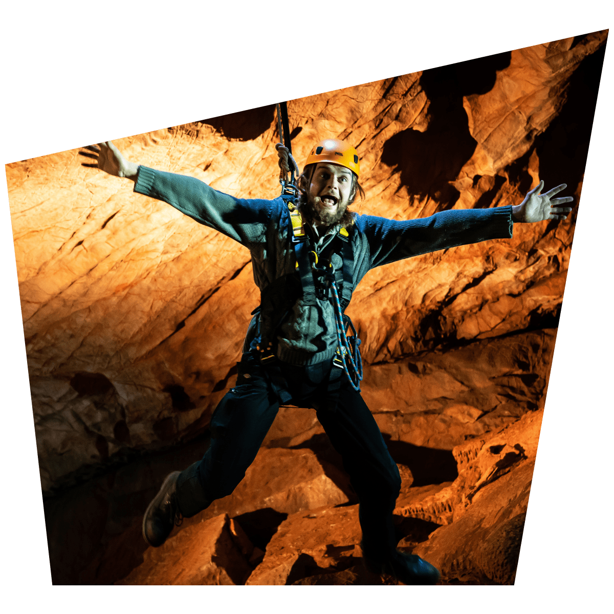 A visitor at Cheddar Gorge, enjoying the Black Cat Freefall activity, as they hang in the cave with a huge grin. The cave is lit with brilliant bright colours to accentuate the cave walls