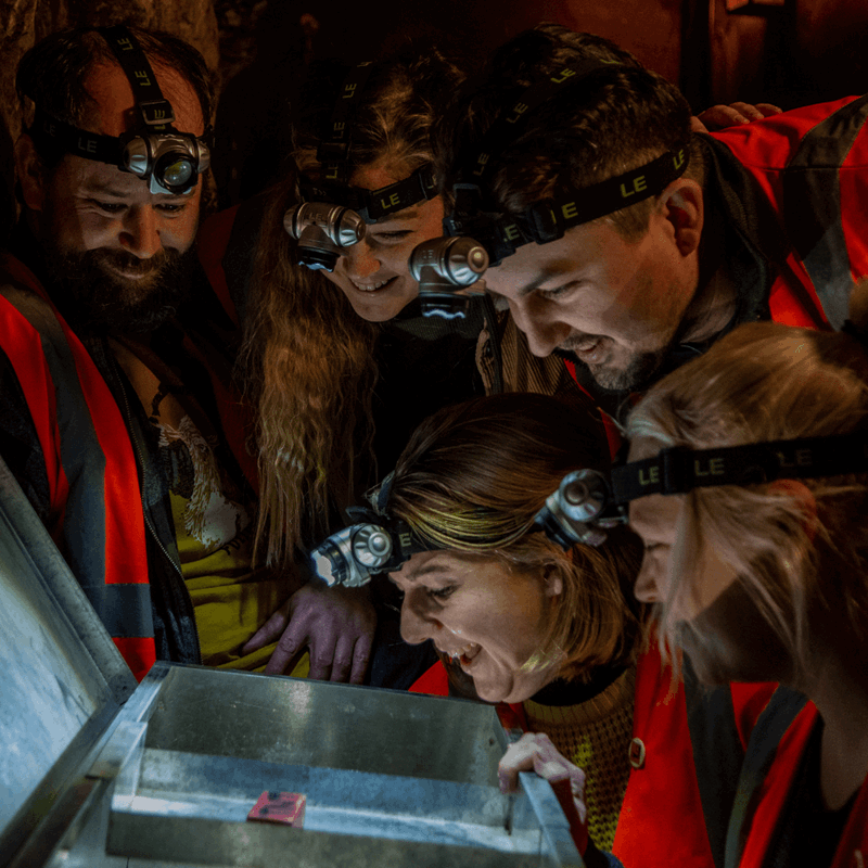 A group of people look at a potential clue in the escape rooms at Cheddar. They gather round a lit up chest looking for answers