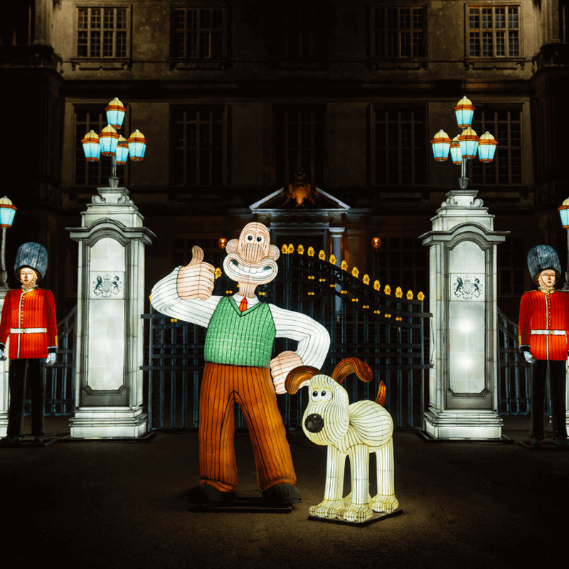Lit up lanterns in the shape of Wallace and Gromit standing in front of fencing guarded by two royal guards in lantern form.