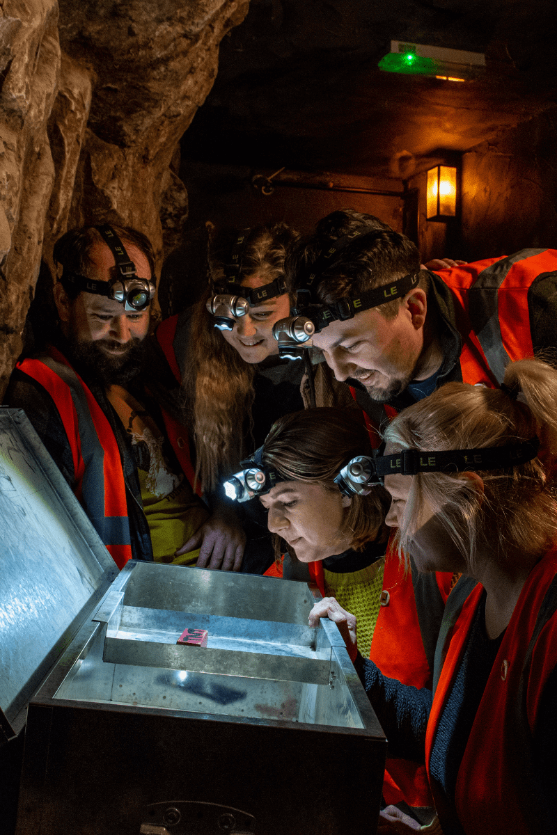 A group of people look at a potential clue in the escape rooms at Cheddar. They gather round a lit up chest looking for answers
