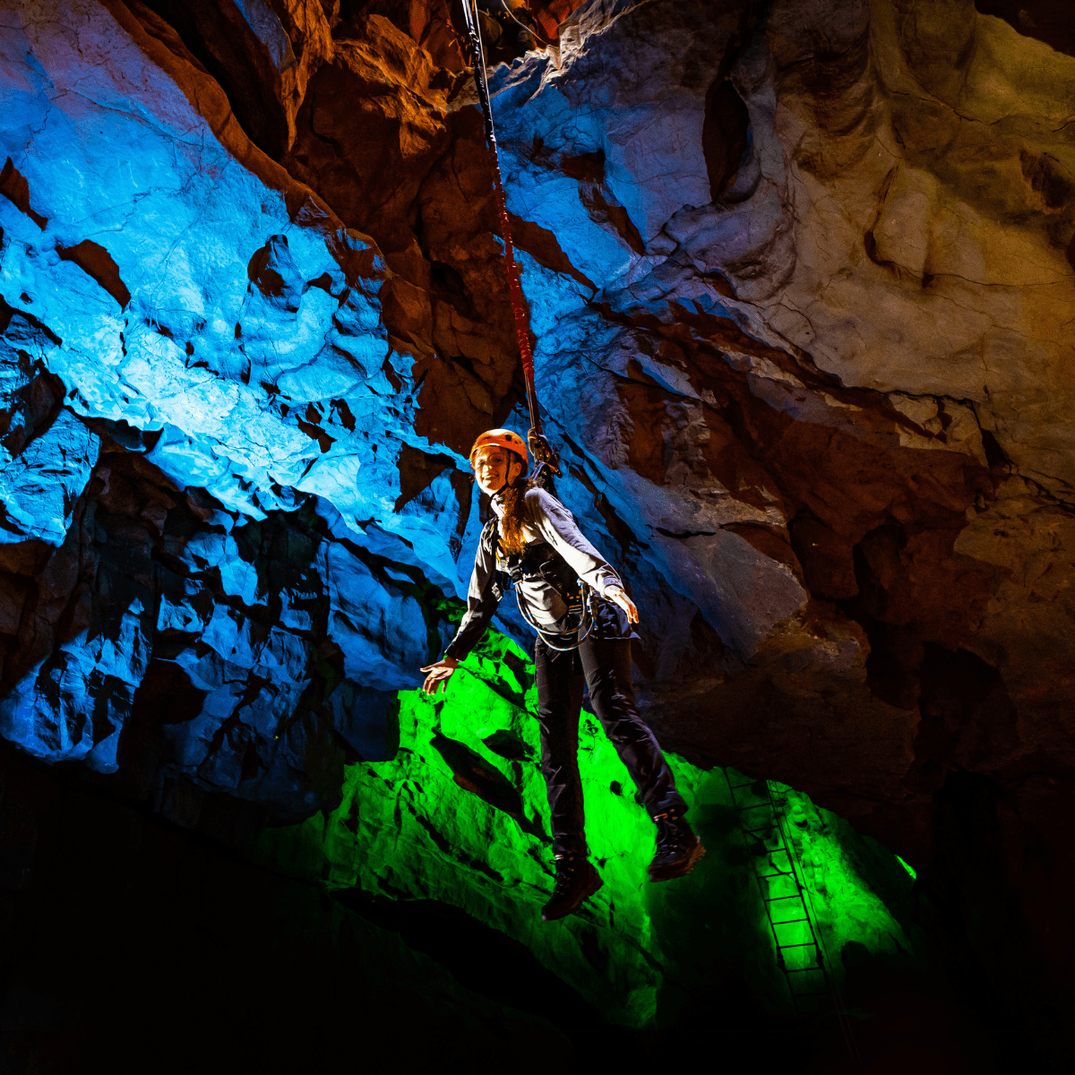 A visitor at Cheddar Gorge, enjoying the Black Cat Freefall activity, as they hang in the cave with a huge grin. The cave is lit with brilliant bright colours to accentuate the cave walls