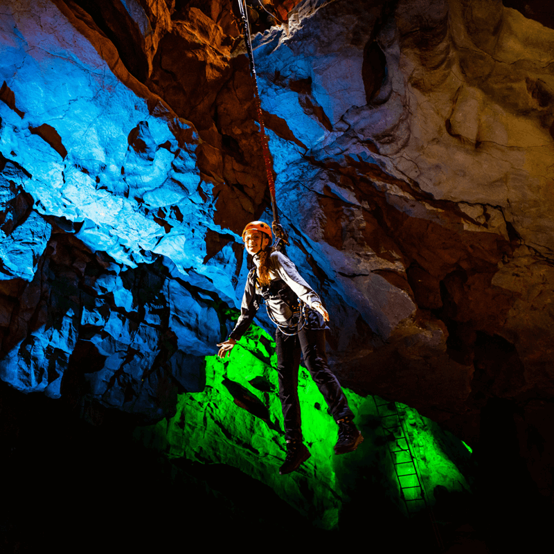 A visitor at Cheddar Gorge, enjoying the Black Cat Freefall activity, as they hang in the cave with a huge grin. The cave is lit with brilliant bright colours to accentuate the cave walls