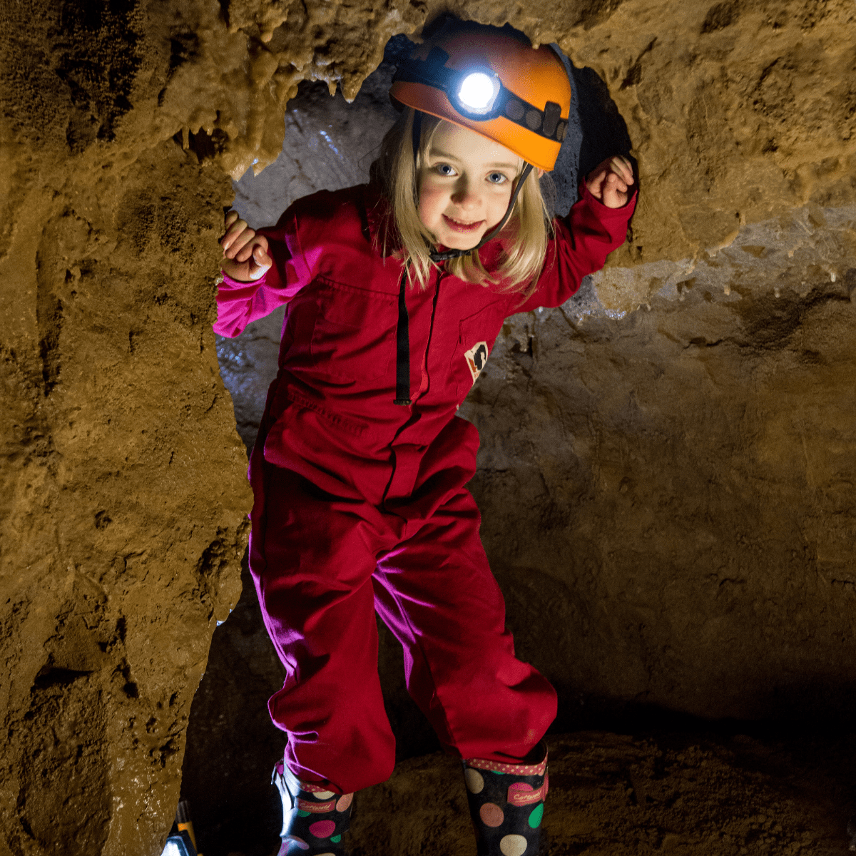 A young girl enjoys taking part in adventure caving, showing her head torch and overalls as she enters through a rock gap