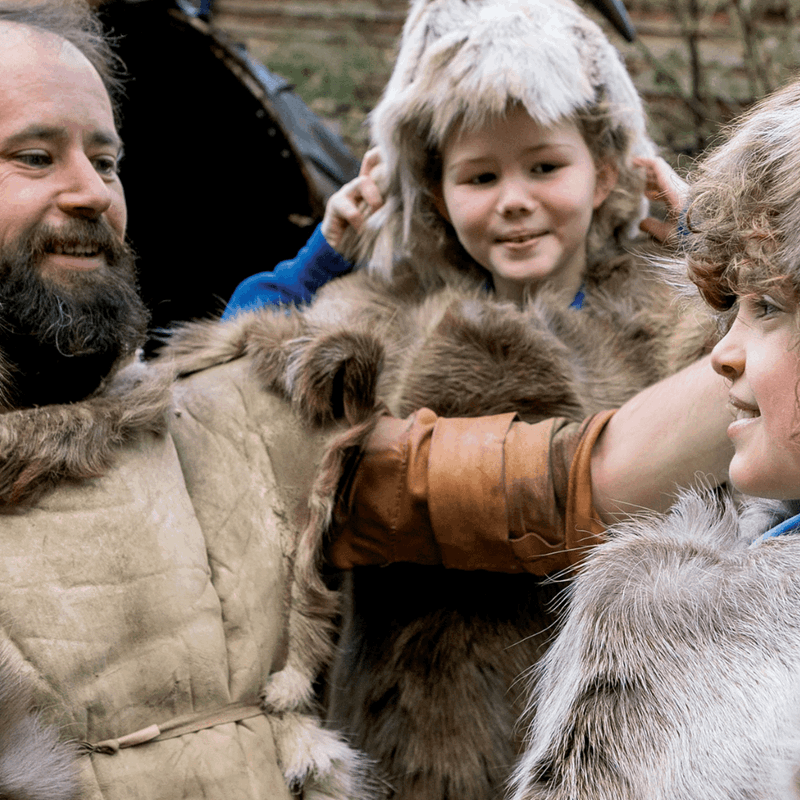 Three children dressed in Stone Age style clothing smile at one another whilst an instructor adjusts a fur hat for one of the children
