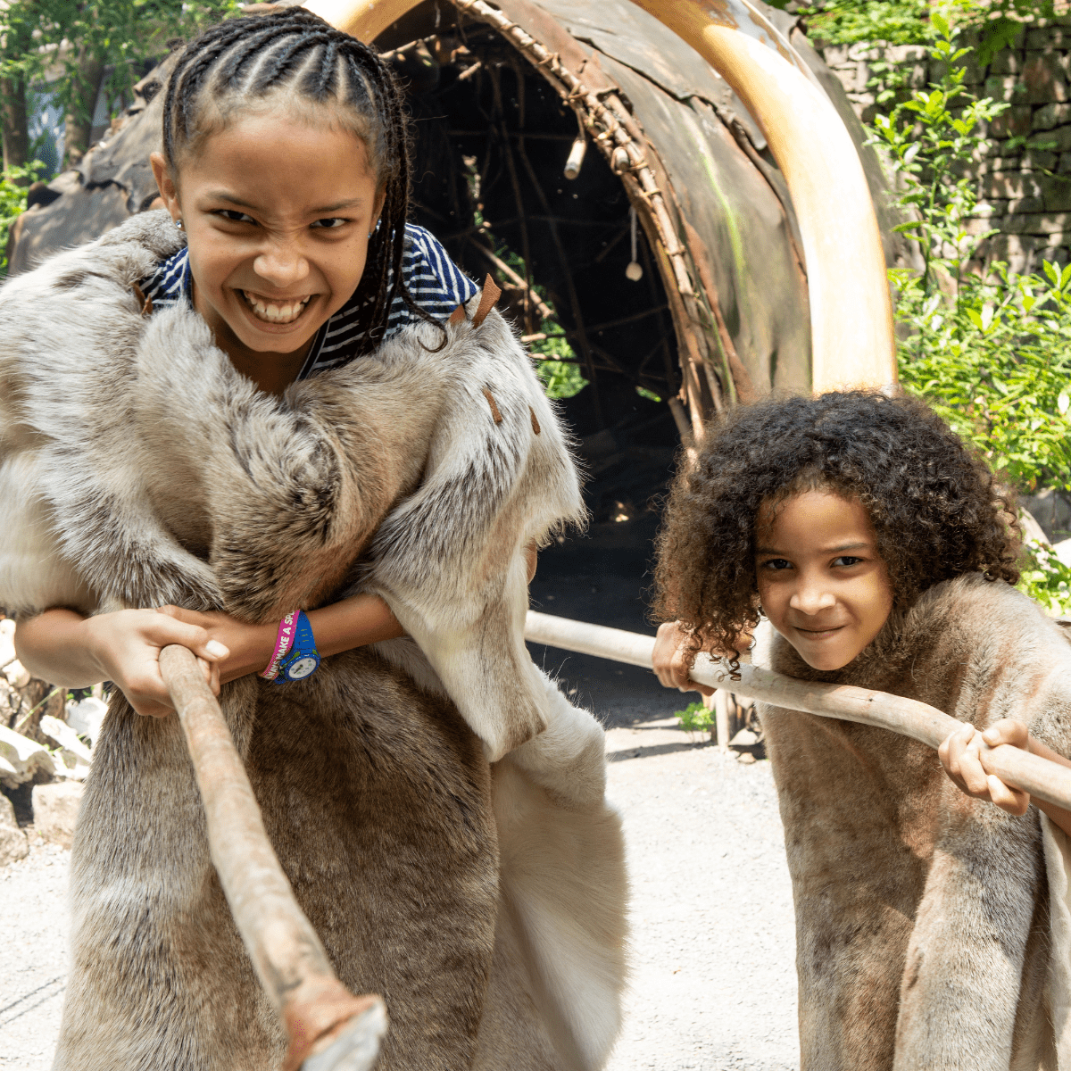 Two girls, dressed in prehistoric clothing hold out wooden staffs in character