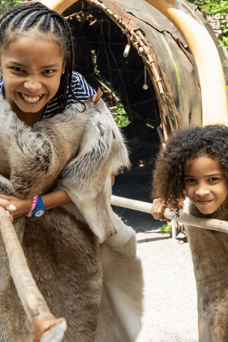 Two girls, dressed in prehistoric clothing hold out wooden staffs in character
