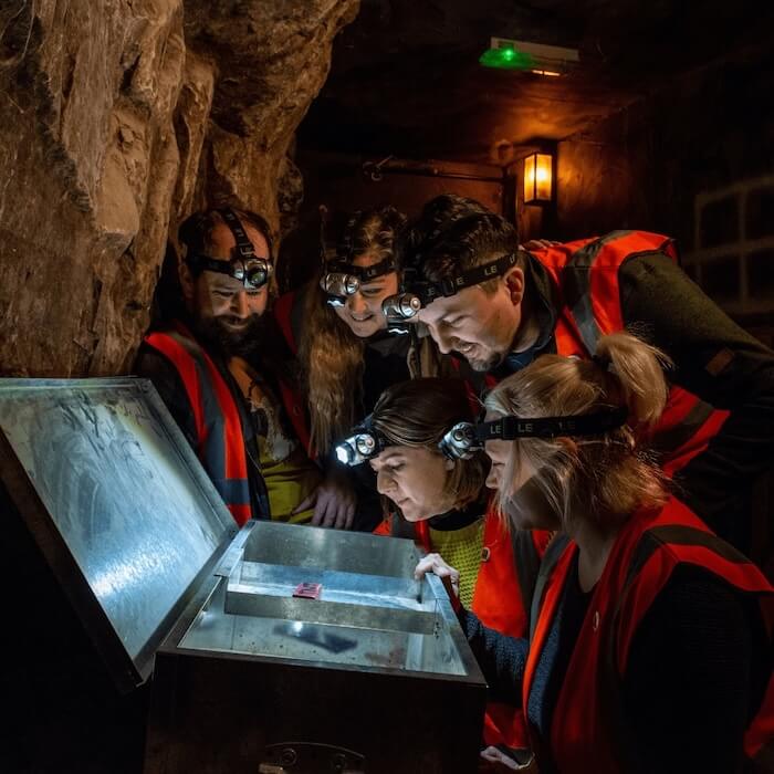 A group of people look at a potential clue in the escape rooms at Cheddar gorge and caves. They gather round a lit up chest looking for answers
