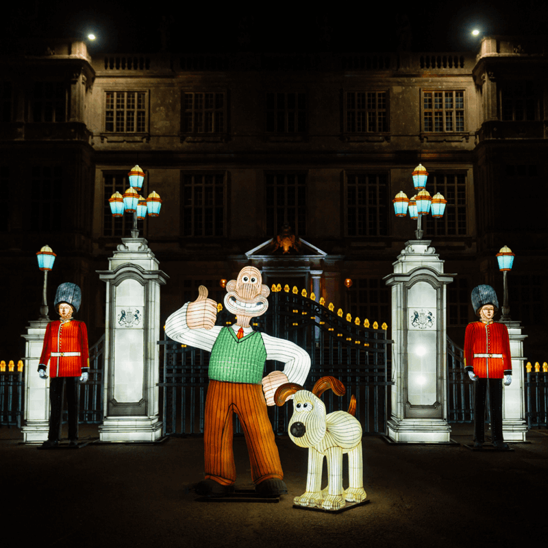 Lit up lanterns in the shape of Wallace and Gromit standing in front of fencing guarded by two royal guards in lantern form.