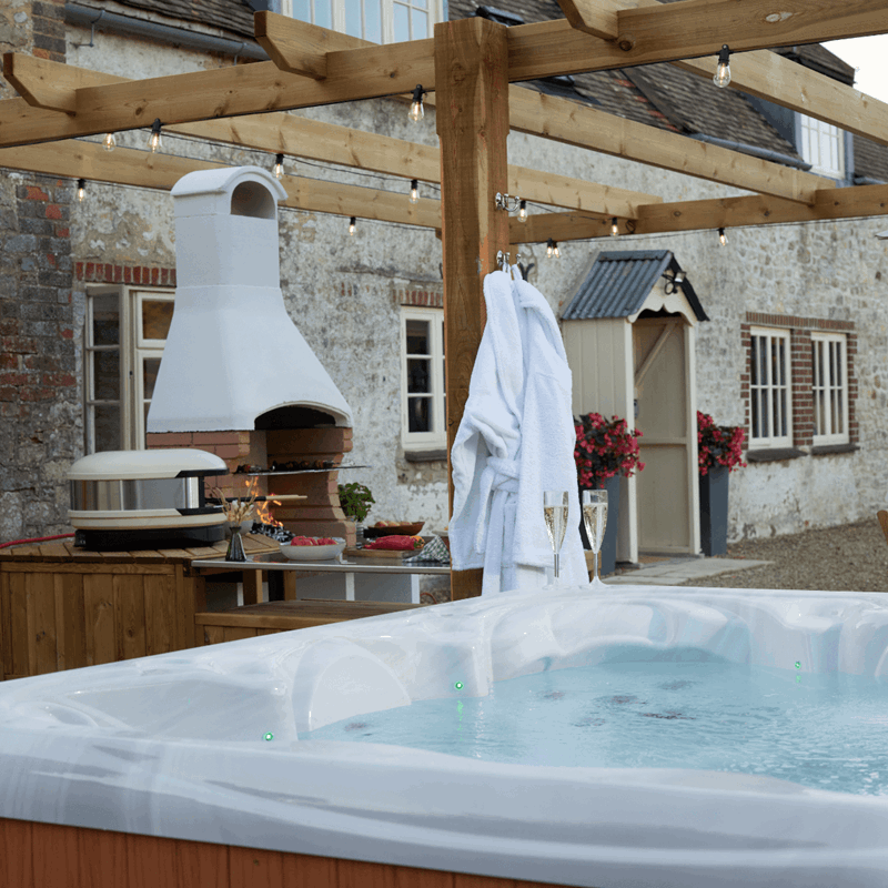 A hot tub with 2 glasses of sparkling wine on the side and robes hanging from a pergola in front of a farmhouse building with pizza oven to the side