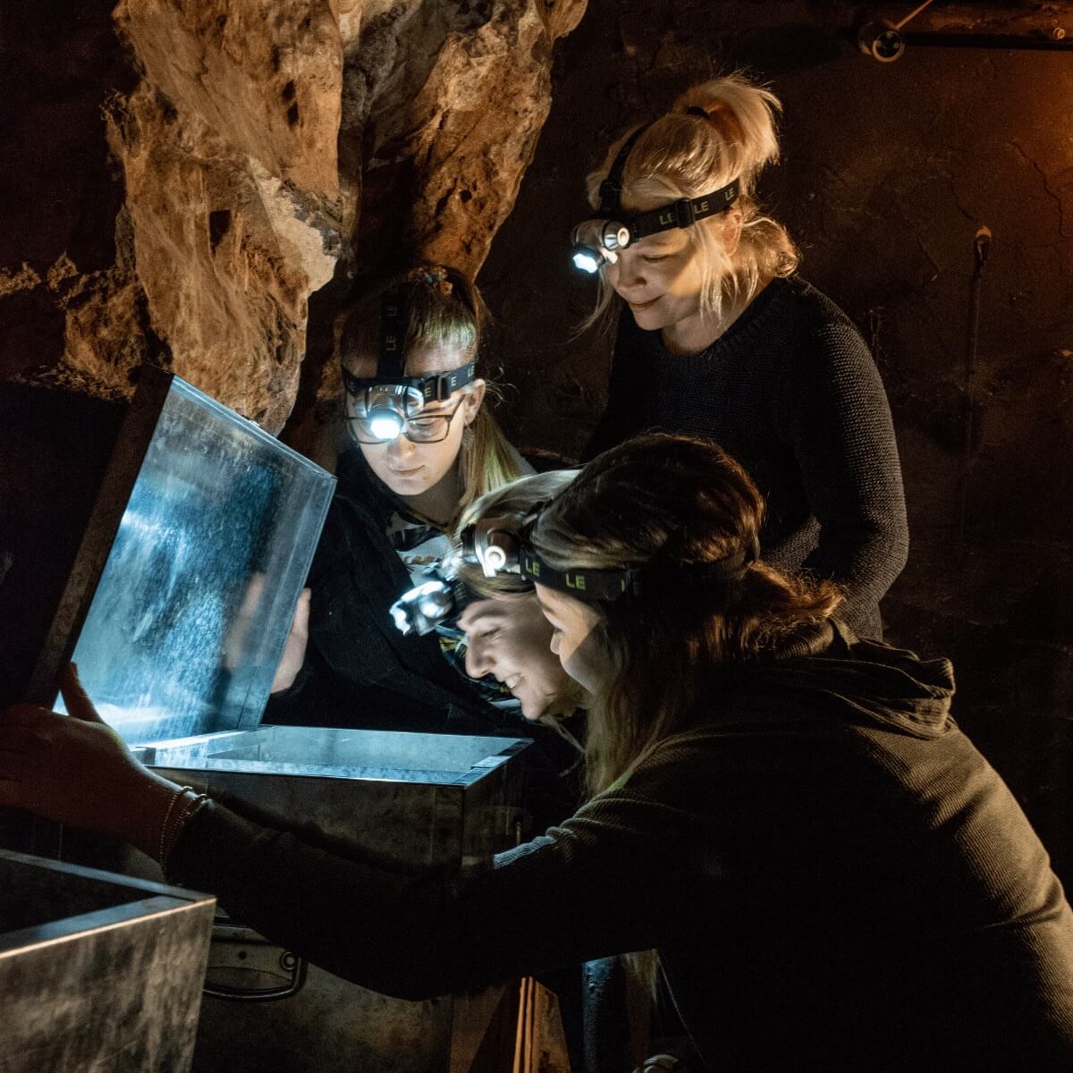 A group of people look at a potential clue in an escape room for family at Cheddar gorge and caves. They gather round a lit up chest looking for answers