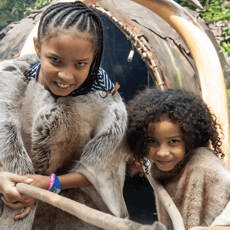 Two girls, dressed in prehistoric clothing hold out wooden staffs in character