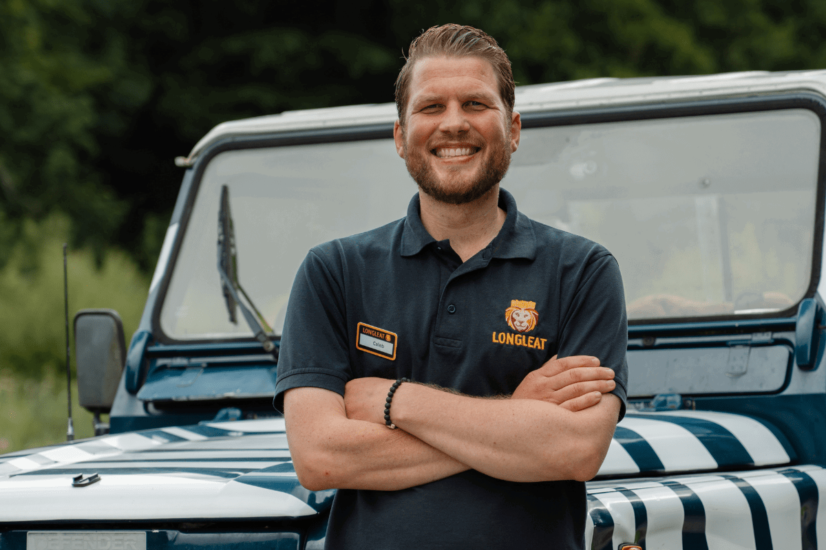 Man stands with his arms folded, smiling, while wearing a blue polo shirt with Longleat Logo. He is leaning against a 4x4 vehicle decorated with zebra stripes.