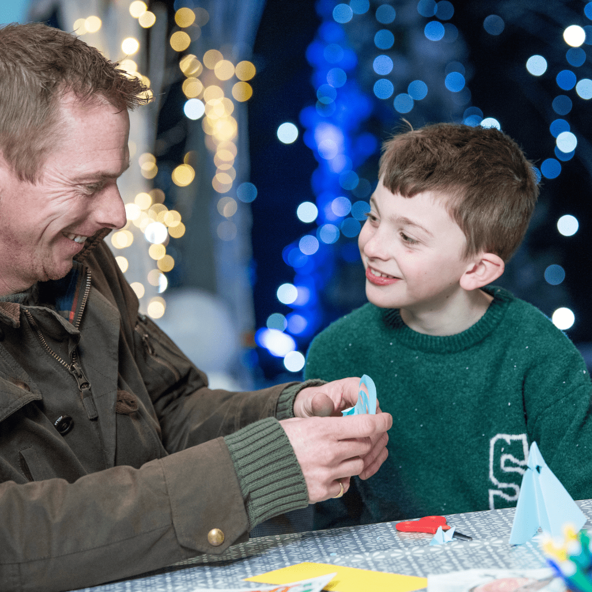 A father and son smiling while doing crafts at a table with Christmas lights shining in the background