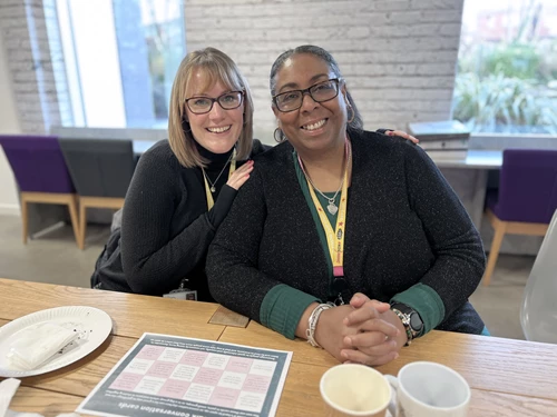 Two people smiling at a table, wearing glasses and ID lanyards.
