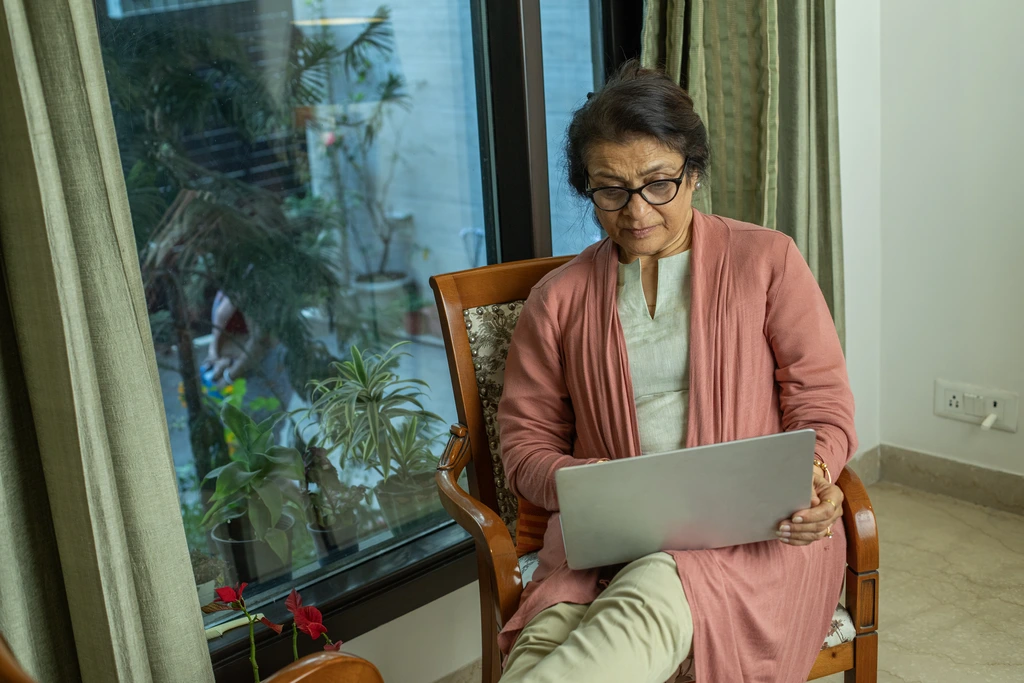 An elderly woman wearing glasses and a pink cardigan sits in a chair by a window, using a laptop