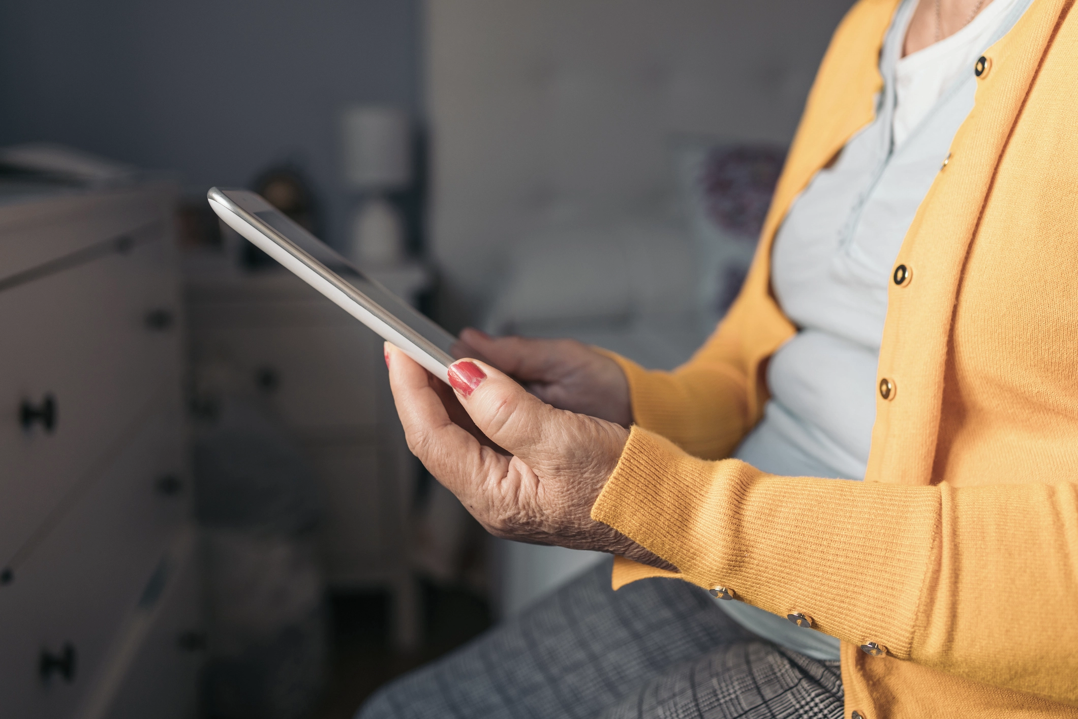 A close up of a woman in a yellow jumper sitting on the side of a bed using her ipad