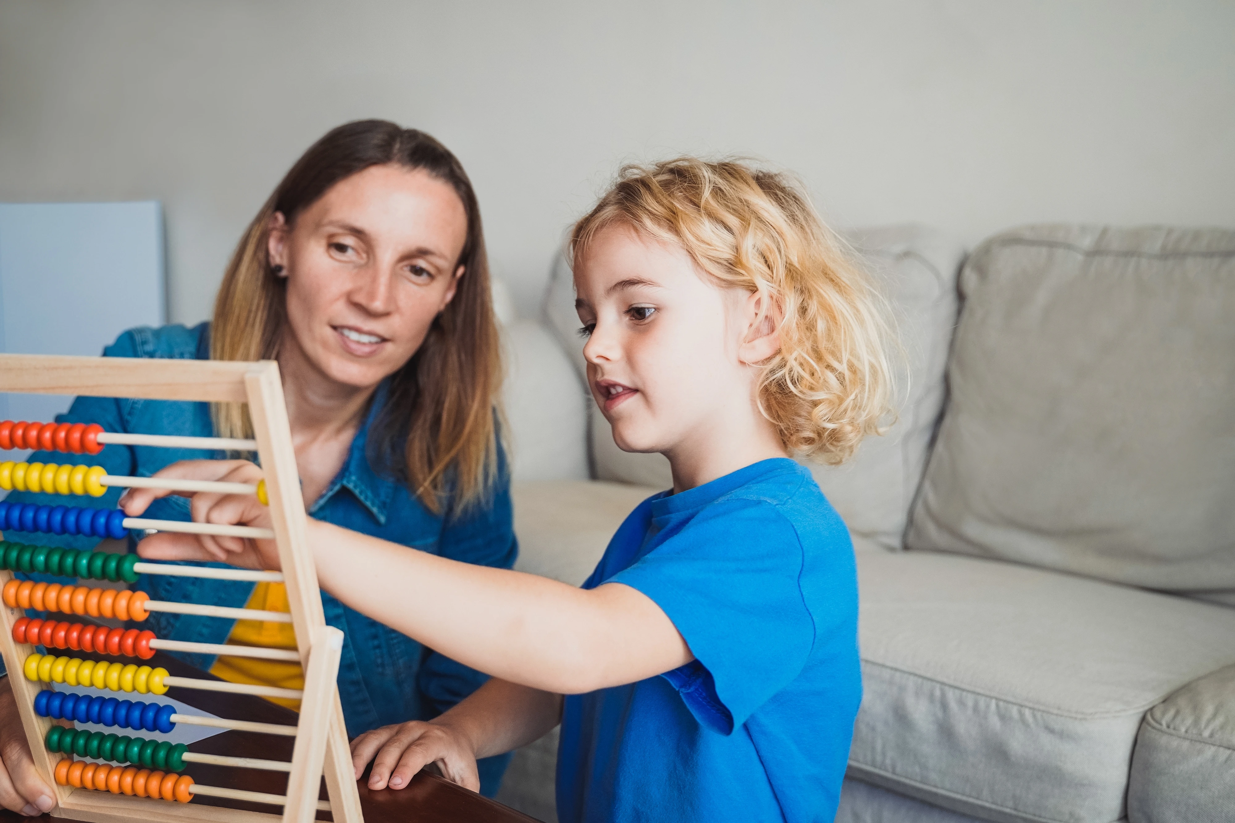 A child with blonde hair and wearing a blue shirt uses an abacus while an adult in a denim shirt watches supportively.