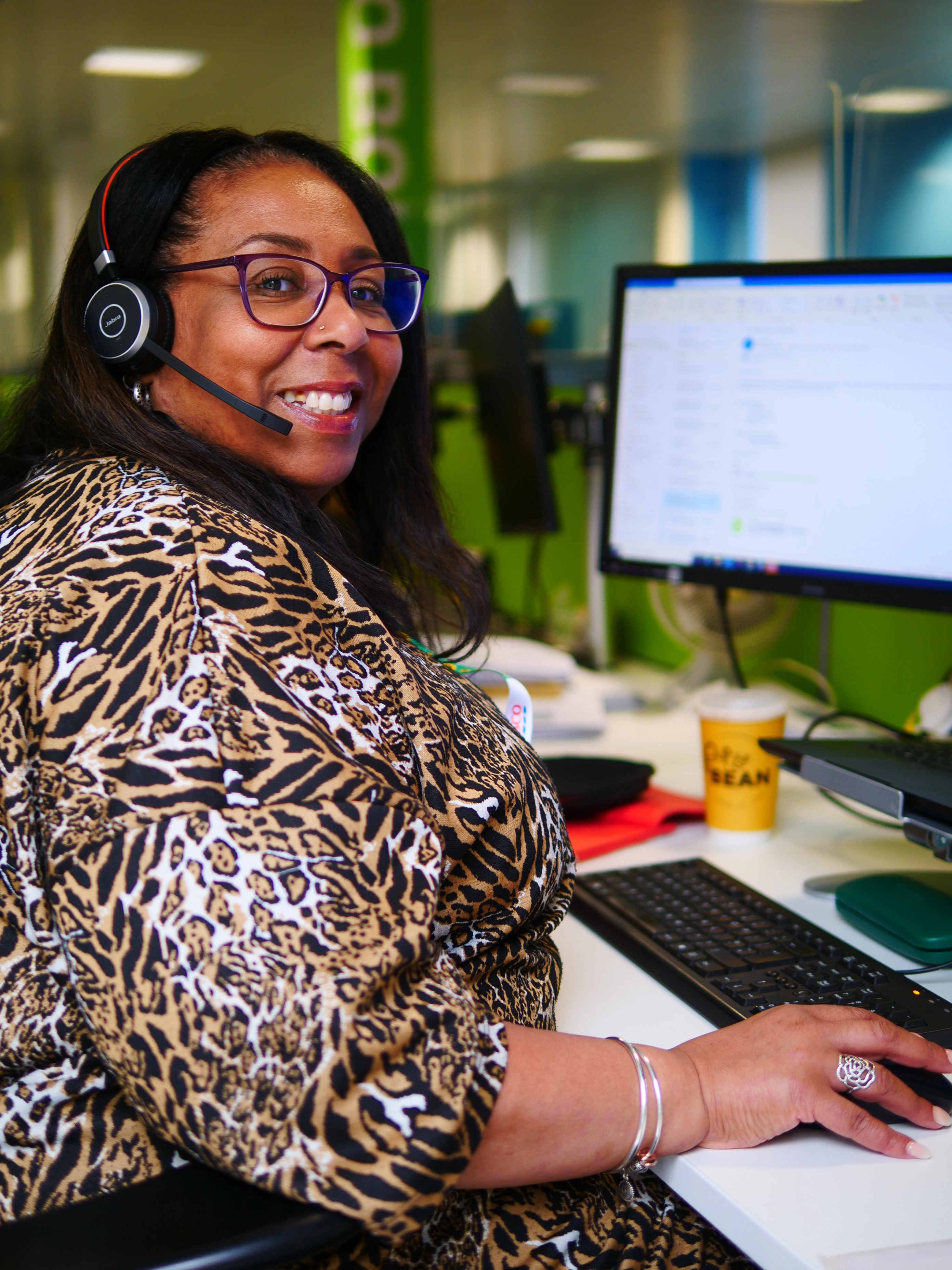 A woman wearing glasses and a headset smiles while seated at a desk with a computer.