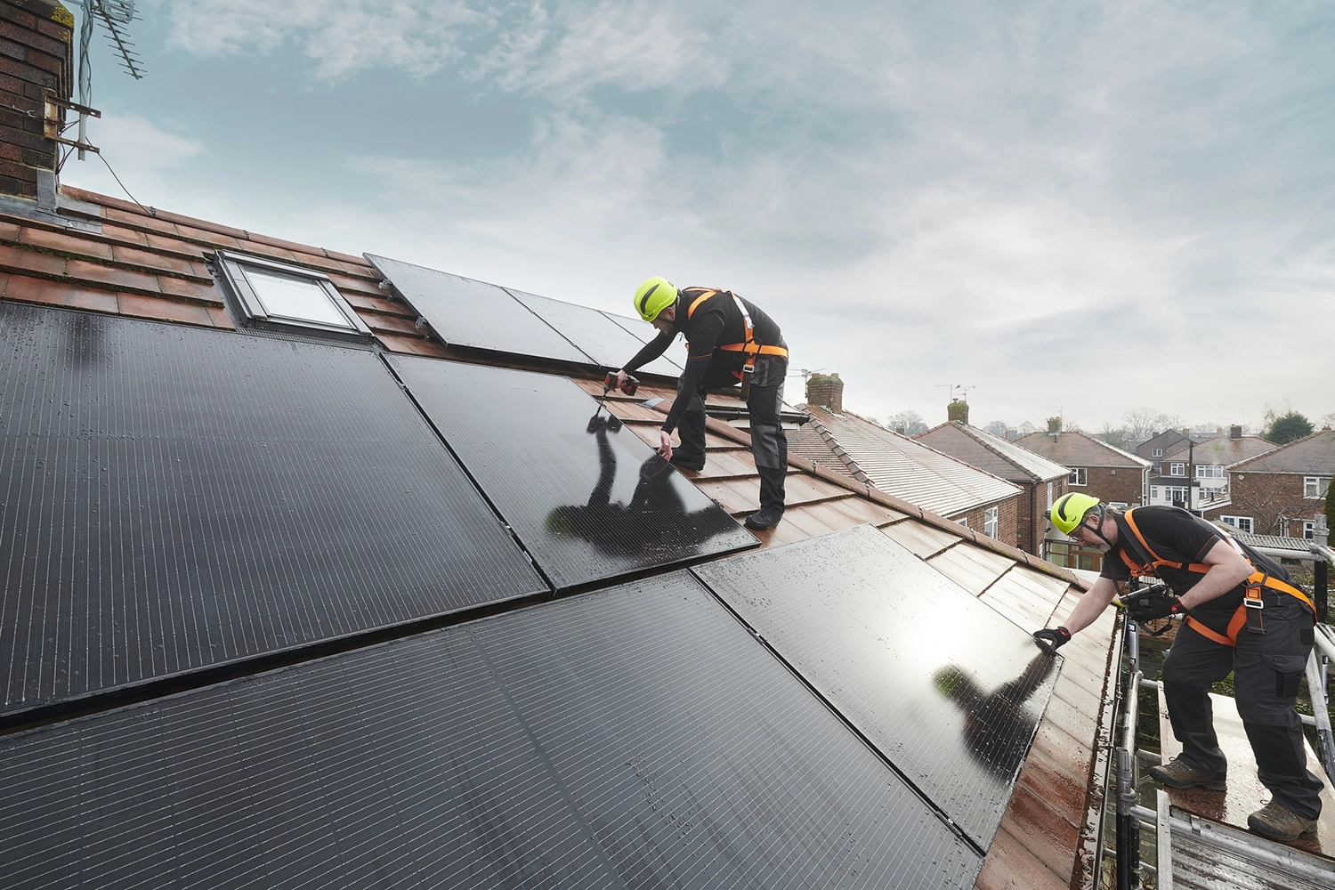 Two workers in safety gear install solar panels on a sloped roof of a house.