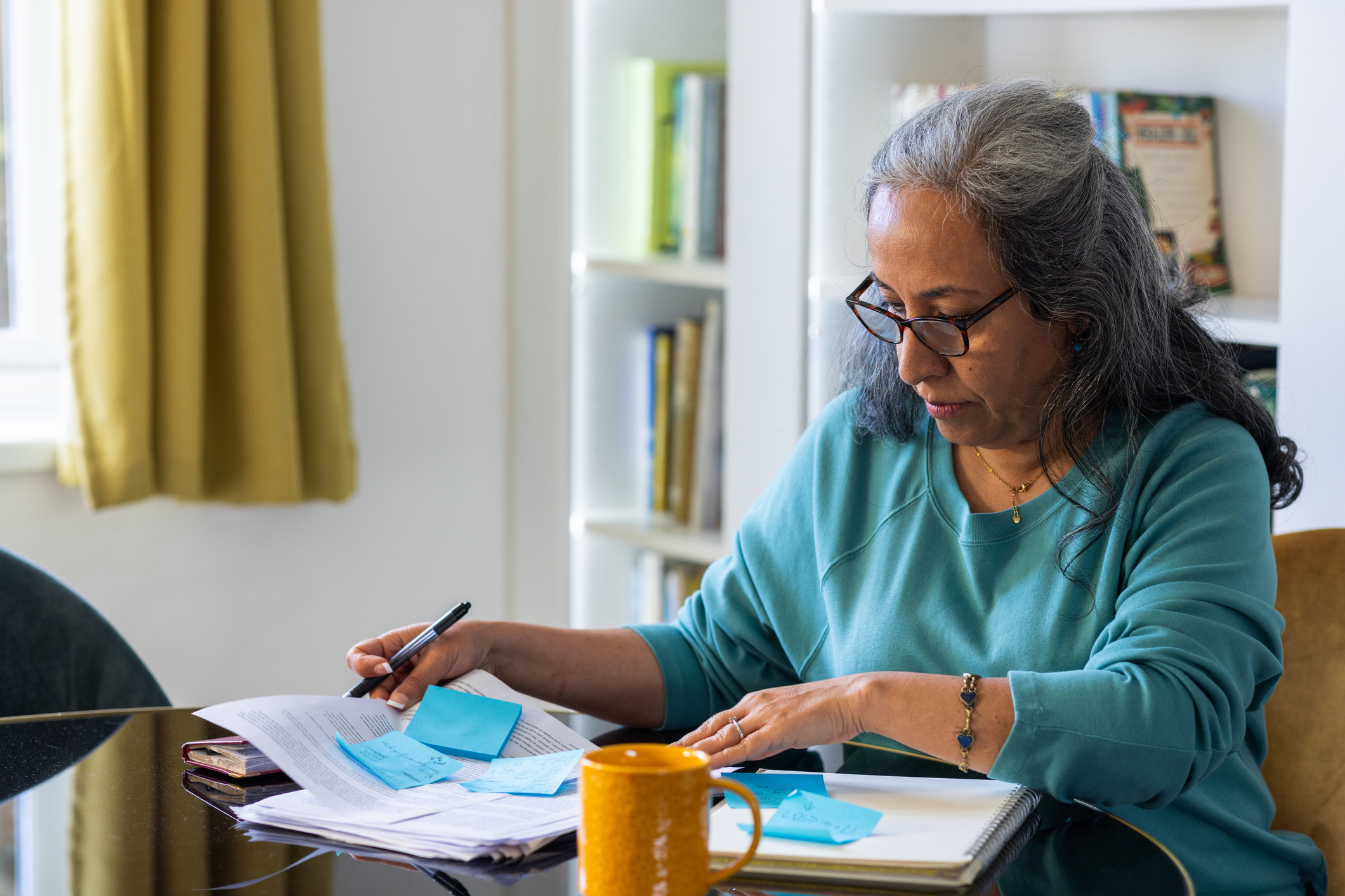 Woman sitting at a table reviewing paperwork with sticky notes and writing with a pen with a yellow mug beside her.