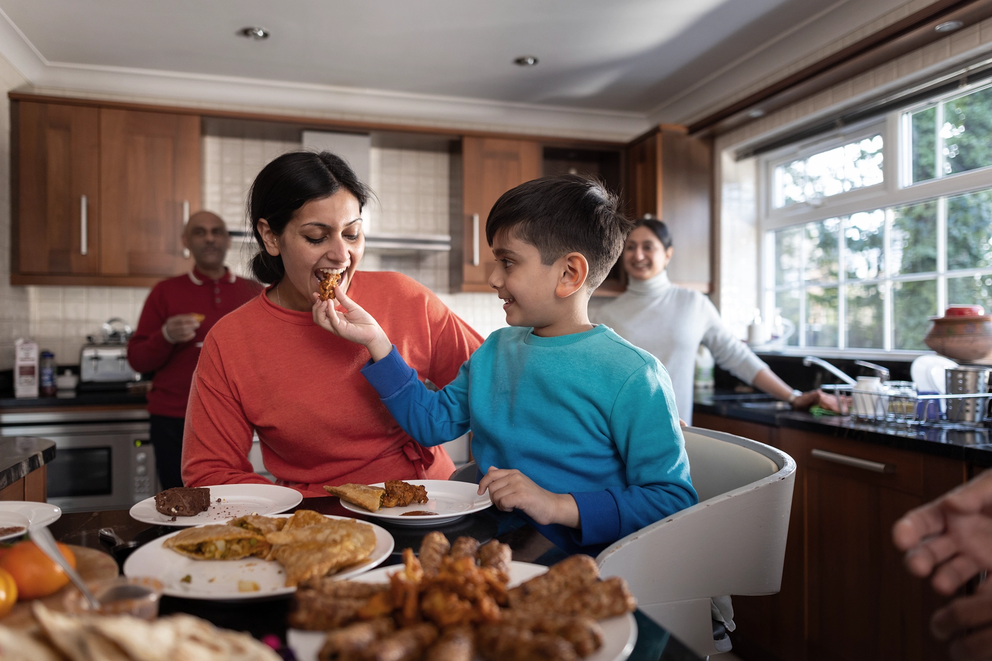 A family of different generations eating food in their kitchen with a boy playfully feeding his mother